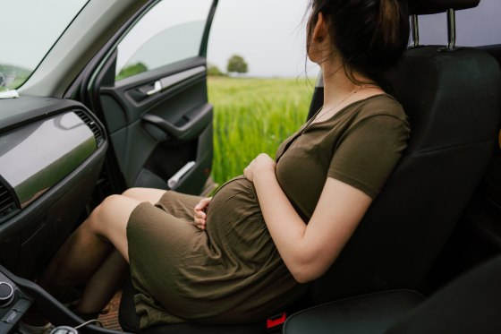 A pregnant woman sitting in a car  in the passenger seat looks out at the view, as seen from the view from the driver's seat