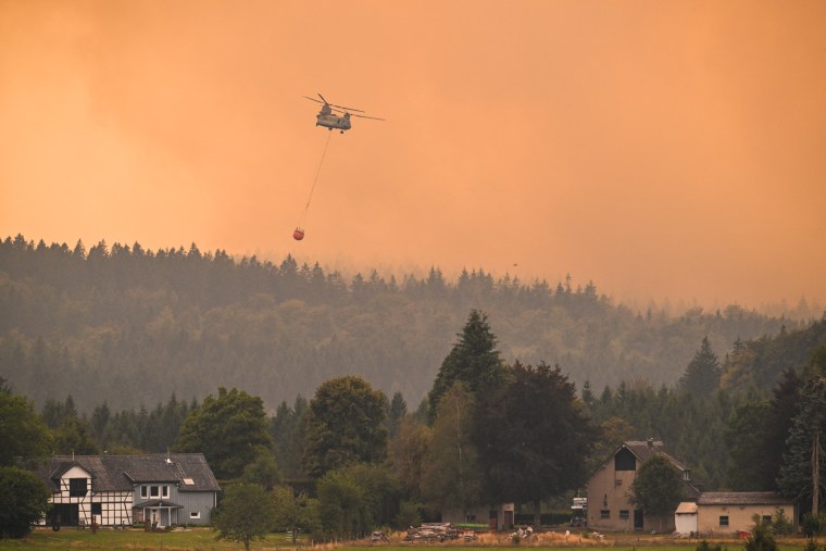 A Royal Netherlands Air Force Boeing CH-47D Chinook carrying a bambi bucket through an orange smokey sky over a forested village