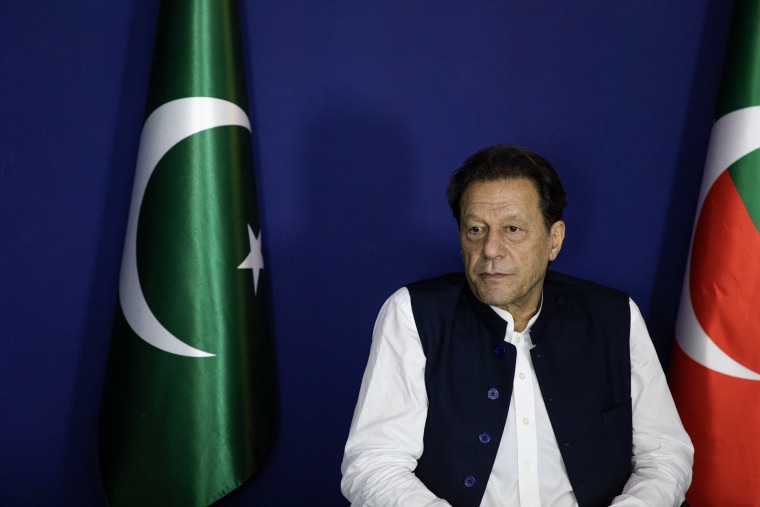 Imran Khan seated between two flags against a blue wall.