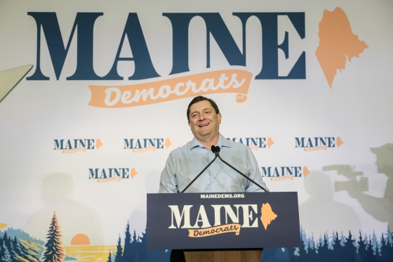 Troy Jackson smiles while standing at a lectern with a "MAINE DEMOCRATS" logo on it.