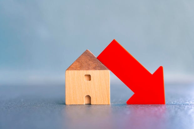 Wooden house model with red down arrow showing decrease in real estate market, home value, or housing demand. Concept of economic recession, property price drop, or mortgage crisis. 